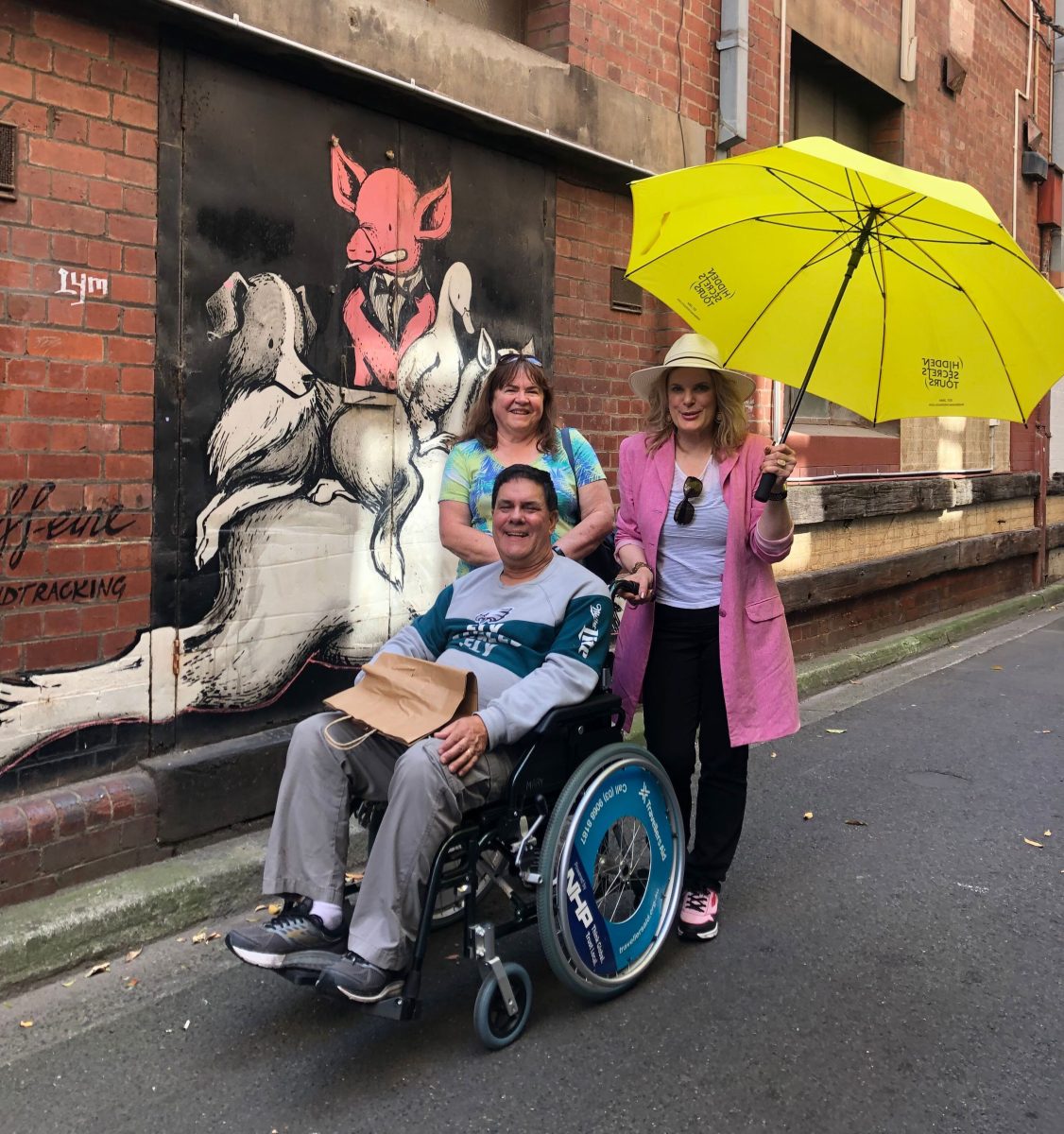 3 people, one in a hired wheelchair observe a lanway that has street art. The tour guide holds a yellow company branded umbrella.