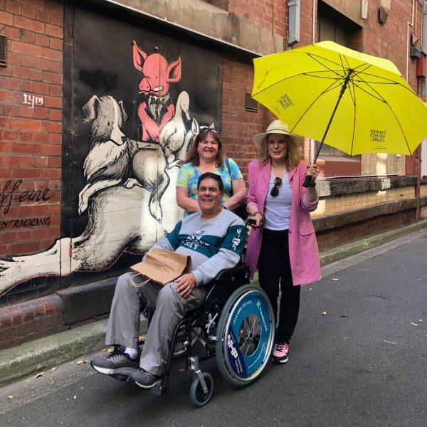 3 people, one in a hired wheelchair observe a lanway that has street art. The tour guide holds a yellow company branded umbrella.